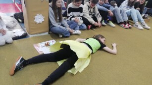 Uczniowie siedzą na podłodze, jedna uczennica leży na podłodze
