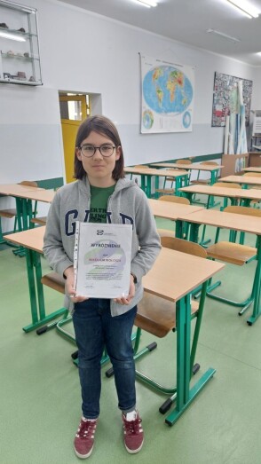 Uczeń w sali lekcyjnej prezentuje dyplom za wyróżnienie w konkursie